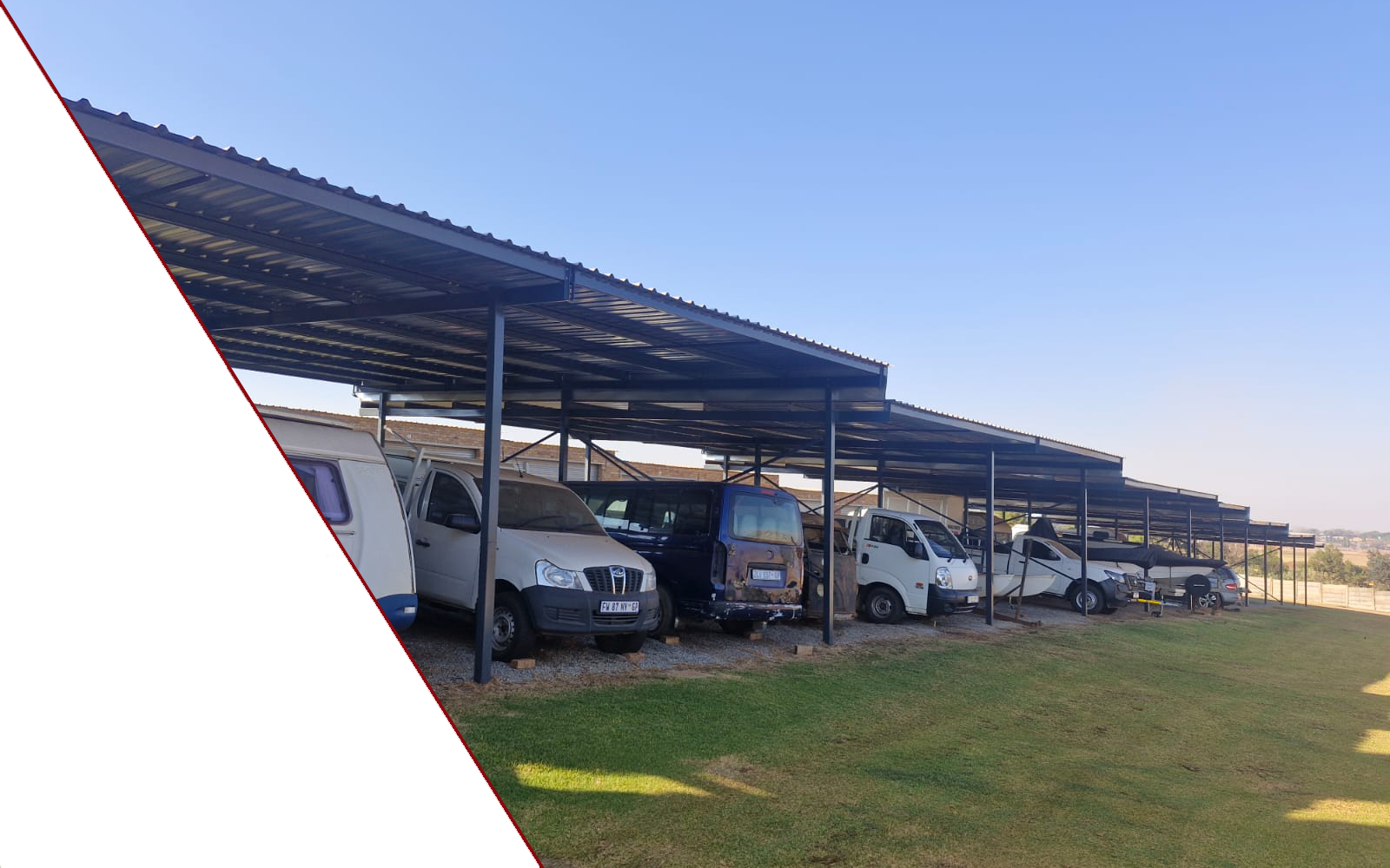 Row of metal carport shelters over parked vans and cars on a grassy area beneath a clear blue sky.