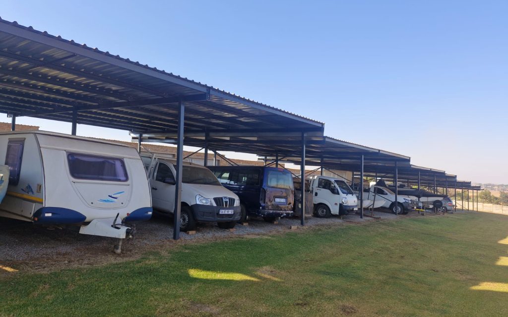 Row of caravans and camper vans parked under metal shade structures in a grassy storage yard.
